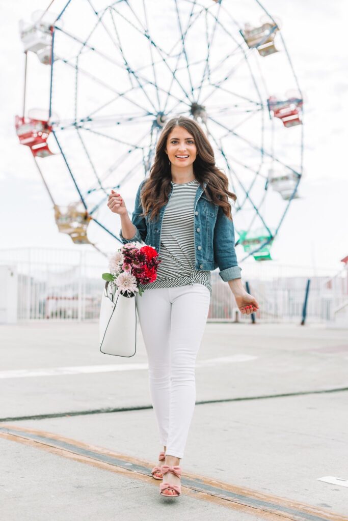 brunette woman in spring jacket and white jeans standing in front of ferris wheel